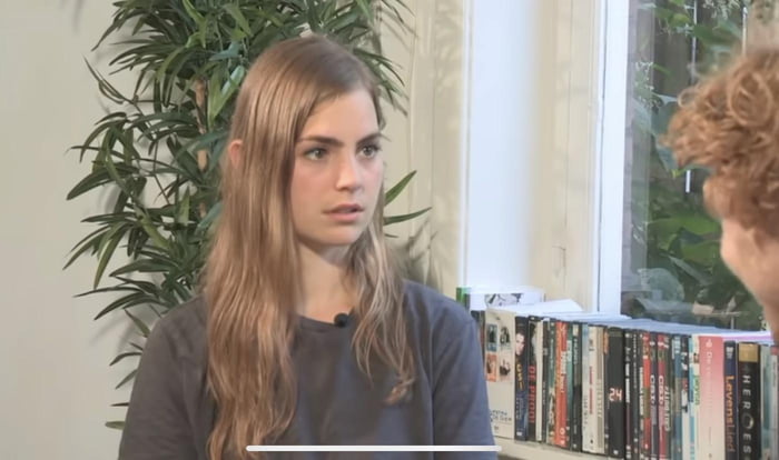 This image captures a young woman with long light brown hair, dressed in a dark grey shirt, sitting in a casual indoor setting. She has a distinct expression of surprise, confusion, and mild concern on her face, appearing to be mid-conversation or interview with another person (only the back of a curly-haired individual's head is visible on the right side of the frame). Behind her, a tall leafy potted plant and a shelf lined with DVD/Blu-ray cases (with partial titles like "HER" and "24" visible) fill the background. This shot is widely used as a reaction meme to convey confusion, disbelief, or being caught off guard by unexpected or absurd statements.