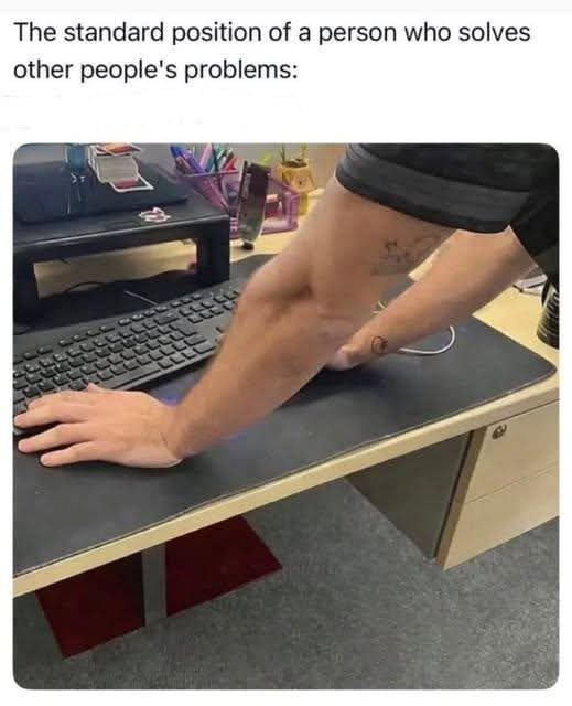 The meme consists of a text caption at the top followed by a photograph. The top text reads 'The standard position of a person who solves other people's problems:'. The photo shows an unidentified person leaning over an office desk in an awkward hunched posture: one of their hands rests on a keyboard on the desk, while their other arm is bent with their elbow tucked under the first arm, pressed against the desk surface. This is a relatable reference to the common uncomfortable position people, especially IT support staff or people helping colleagues with tech issues, take when troubleshooting someone else's computer quickly without sitting down at their desk.
