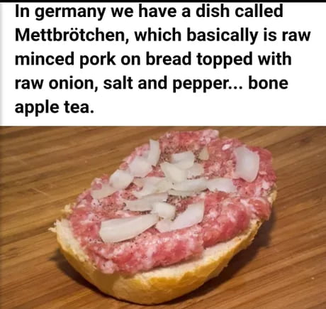The meme consists of top text introducing the traditional German savory dish Mettbrötchen, and a photo of the dish below the text. The text explains that Mettbrötchen is a bread roll topped with raw minced pork, raw onion, salt and pepper, and ends with the humorous intentional phonetic misspelling of the French phrase 'bon appétit' as 'bone apple tea', a popular internet meme phrasing. The photo shows a Mettbrötchen sitting on a wooden cutting board, clearly displaying the layer of raw minced pork, scattered raw onion slices, and seasoning on top of the bread. The joke plays on both the unfamiliarity of raw pork dishes to many non-German audiences and the widely shared silly misspelling of 'bon appétit'.