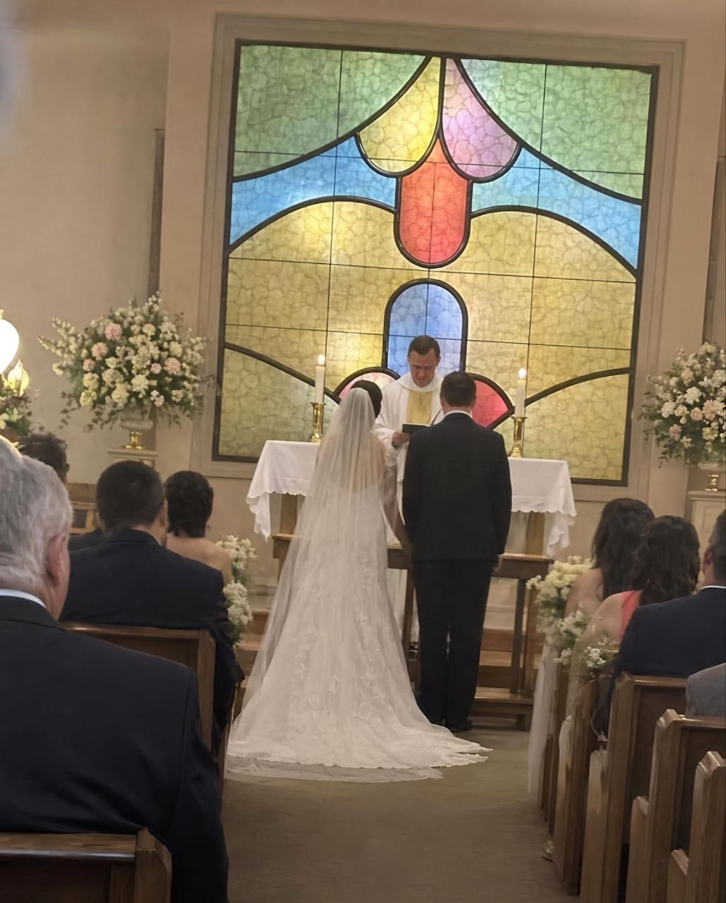 This is a candid photograph taken from the audience seating during a wedding ceremony held inside a church. A bride wearing a long white lace wedding gown and veil, and a groom in a black suit stand at the altar facing a priest who is officiating the service. The large stained glass window positioned directly behind the altar has a color-blocked pattern that unintentionally perfectly matches the shape and design of a crewmate character from the popular video game *Among Us*, creating a humorous unintended visual joke for viewers familiar with the game. Guests are seated in wooden pews on both sides of the aisle watching the ceremony, with white and pale pink flower arrangements decorating the altar and aisle edges.