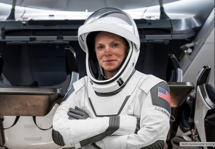A photograph of astronaut Zena Cardman wearing a white spacesuit with an American flag patch on the right shoulder and the name 'Z. Cardman' printed on the chest. She stands with her arms crossed inside what appears to be the interior of a spacecraft, with metallic seats and equipment visible in the background. The image is accompanied by a speculative comment from 9Gag suggesting Crew-11 is returning early due to a 'medical condition,' with the claim that Cardman is pregnant.