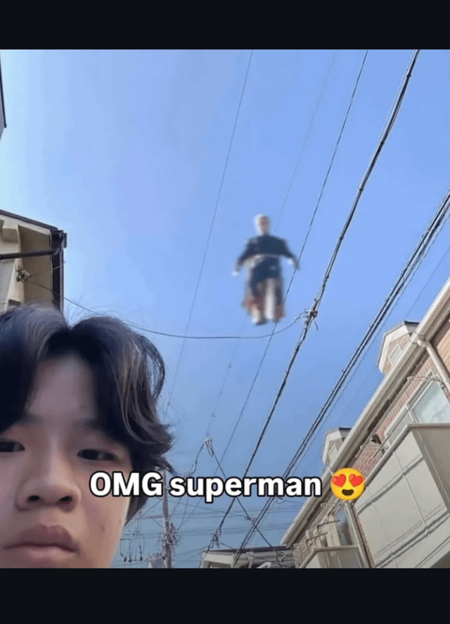A selfie taken in an alley with residential buildings on both sides. The sky is clear blue with multiple power lines crisscrossing overhead. In the center of the sky, a small figure is suspended between the power lines, appearing to float. At the bottom of the image, white overlaid text reads 'OMG superman 😍' with a heart-eyed emoji, humorously implying the floating figure is Superman.