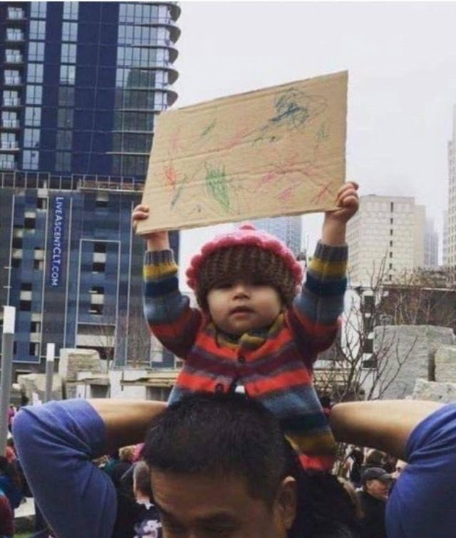 This candid photograph captures a young toddler wearing a fuzzy pink knit hat and a colorful striped fleece jacket, perched on the shoulders of an adult man at an outdoor public gathering, likely a protest or rally, in a downtown city area with modern high-rise buildings visible in the background. The toddler holds a blank piece of cardboard aloft, which is covered only in colorful, unstructured child scribbles rather than the text-based slogans typical of protest signs, creating a lighthearted, humorous juxtaposition. The toddler wears a deadpan, unimpressed neutral expression, adding to the comedic value of the image, which is commonly used as a reaction meme.