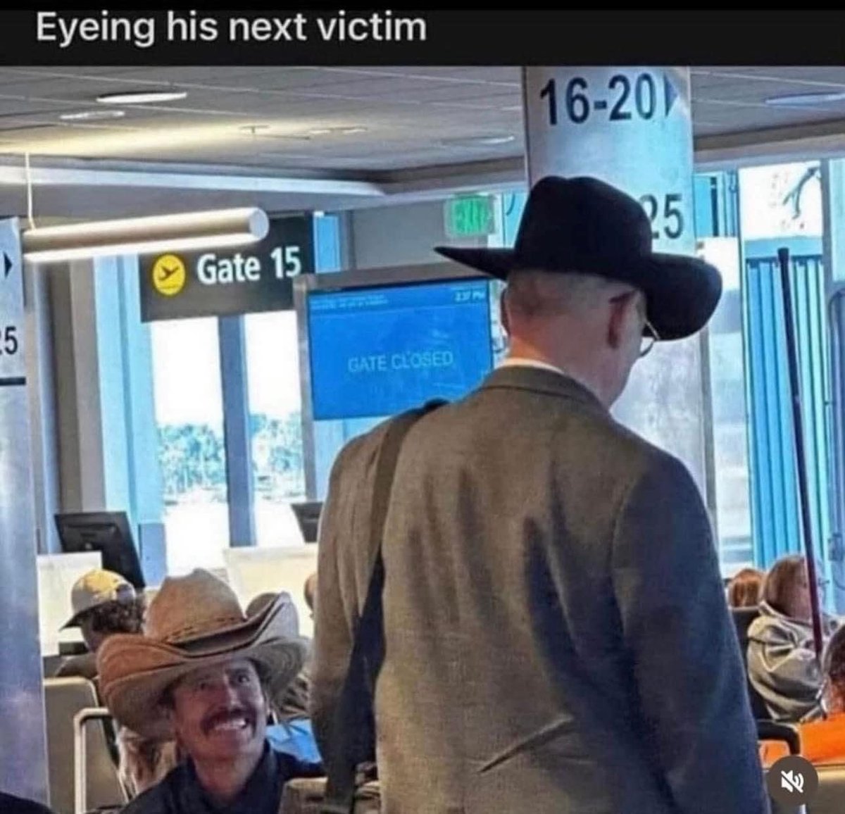 This meme features a candid photograph taken inside a commercial airport terminal, near gate 15 where a blue digital sign displays the text 'GATE CLOSED'. In the foreground, an older man seen from the back wears a black cowboy hat, grey suit jacket, and glasses, standing and facing towards the gate area. Seated behind him to the left is another man wearing a distressed tan cowboy hat, with a prominent dark mustache, who is grinning widely and looking directly at the back of the standing man as if plotting something. The humorous text overlay at the very top of the image reads 'Eyeing his next victim', framing the seated cowboy's amused expression as a scheming, playful threat towards the unsuspecting standing man. Additional airport signage indicating gates 16-20 and 25, overhead lighting, and other waiting passengers are visible in the background.