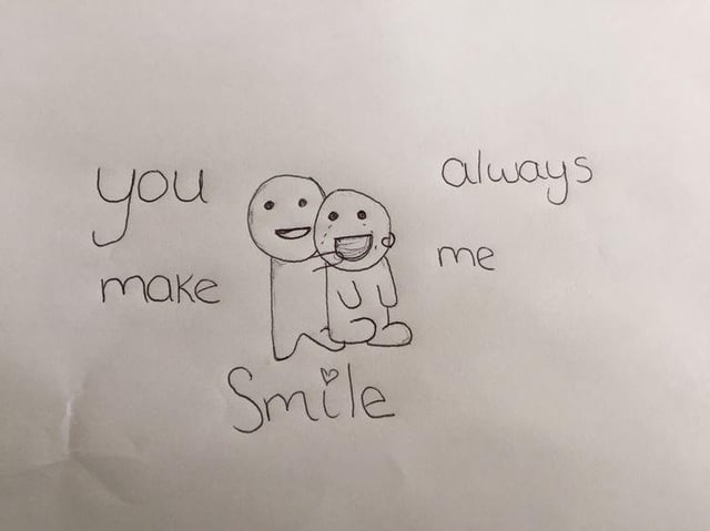 A hand-drawn sketch on white paper featuring two stick figures. The left figure stands and hugs the right seated figure. Both have smiling expressions: the left figure has a simple closed smile, while the right figure has an open-mouthed grin. Surrounding the figures is handwritten text arranged to form the phrase 'you make me always Smile': 'you make' is written to the left of the standing figure, 'always me' to the right of the seated figure, and 'Smile' (with a small heart dotting the 'i') below the figures. The paper has a faint crease visible on the left side, indicating minor wear.