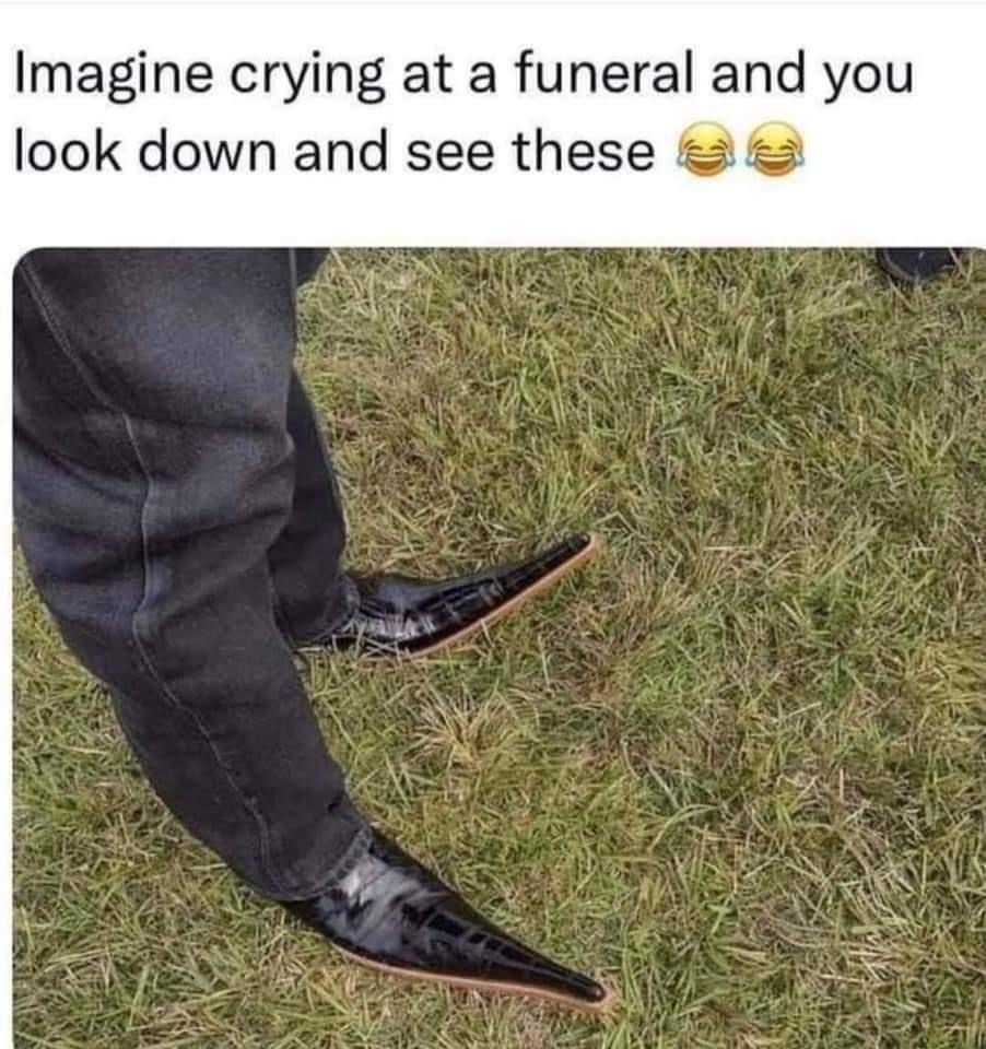 The meme is composed of text at the top and a photograph below. The top text reads 'Imagine crying at a funeral and you look down and see these' paired with two laughing with tears emojis. The photo below shows the lower legs of a person standing on a grassy surface, wearing dark trousers and extremely long, sharply pointed, shiny black dress shoes with an absurdly exaggerated, over-the-top design. The humor comes from the sharp juxtaposition between the somber, serious context of a funeral where people are expected to grieve, and the ridiculous, out-of-place appearance of these shoes, which would be so comically inappropriate that they would break the serious mood even when someone is trying to cry.