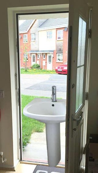 The image depicts a domestic scene where a white pedestal sink is positioned in the middle of an open glass door, completely blocking entry or exit. The door leads to an outdoor area featuring row houses with brick exteriors, a red car parked on the street, green grass, and a concrete path. The indoor side shows part of a hallway with a doormat near the door and some indistinct items along the wall. The sink's placement creates a confusing and impractical obstacle in the doorway.