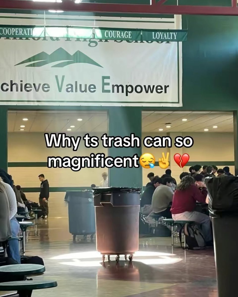 This is a humorous meme photo taken inside a high school cafeteria or common area. A standard large wheeled plastic trash can is positioned in the center of the frame, with a bright beam of overhead light shining directly on it, casting a dramatic glow that makes the mundane object look unintentionally important and grand. In the background, groups of high school students are seated at cafeteria tables socializing or eating, with a large school values banner hanging above the doorway, displaying the words 'COOPERATION, COURAGE, LOYALTY' and 'Achieve Value Empower' alongside a mountain logo. Overlaid on the center of the image is text that reads 'Why ts trash can so magnificent' followed by a sleepy drooling face emoji, a peace hand emoji, and a broken heart emoji, expressing exaggerated, playful admiration for the accidentally majestic-looking trash can. The joke comes from the absurd contrast between the dramatic lighting on a boring everyday object and the overly emotional praise in the text.