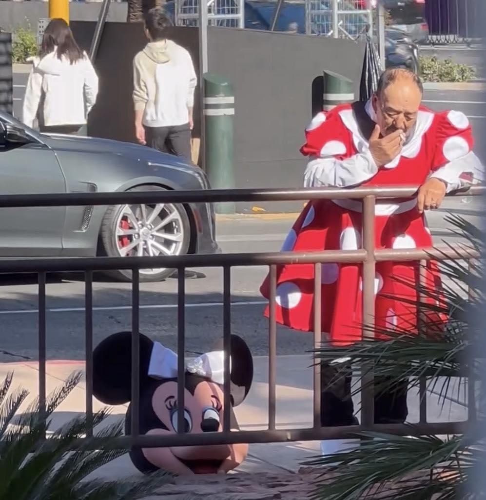 The image is a candid street photograph featuring an older bald man wearing only the body section of a Minnie Mouse costume: a bright red dress with large white polka dots and puffy white sleeves. He is leaning against a brown metal pedestrian railing on a sidewalk, holding a cigarette to his mouth as he takes a smoke break. The full, cartoonish Minnie Mouse headpiece with large black ears, a white bow, and smiling face sits discarded on the sidewalk at the base of the railing in front of him. In the background, a silver sedan is parked at the curb, two pedestrians walk away from the camera past dark barrier walls, and street bollards are visible. The humor of the image stems from the jarring, relatable contrast between the wholesome, family-friendly public persona associated with the Disney character costume and the mundane, tired, unglamorous reality of the worker wearing it during a work break.