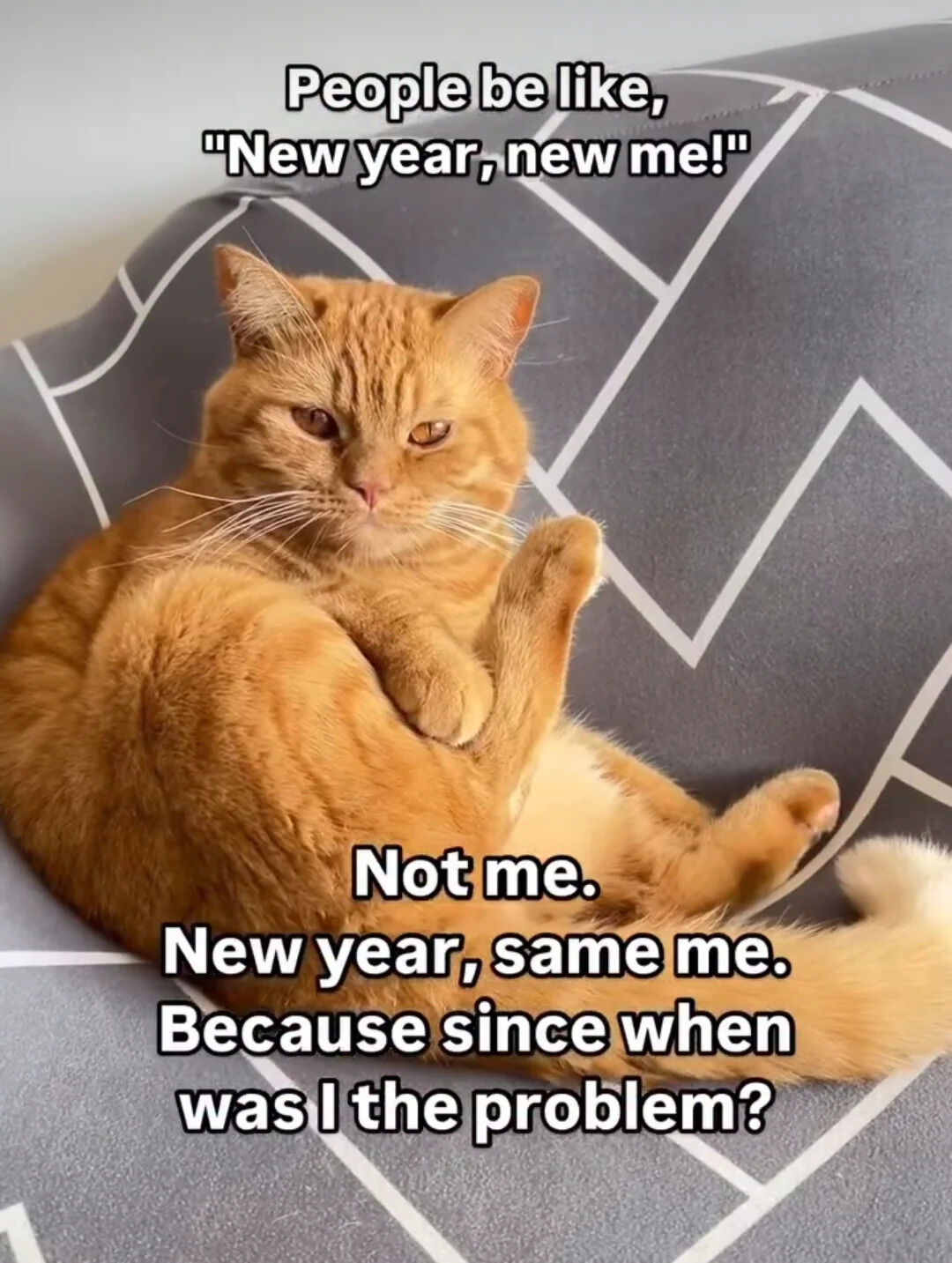 A photograph of an orange tabby cat lying curled up on a gray cushion with white geometric patterns. The cat has a relaxed, slightly indifferent expression with half-lidded eyes and one paw raised. Overlaid text humorously contrasts societal New Year's resolution culture: top text reads 'People be like, "New year, new me!"' and bottom text (framed as the cat's perspective) reads 'Not me. New year, same me. Because since when was I the problem?'