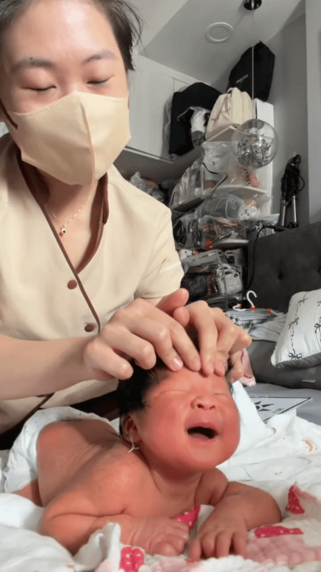 The image captures a real-life scene where a woman wearing a beige face mask and beige uniform is gently giving a head massage to a newborn baby. The newborn is lying prone on a soft blanket with pink polka dot details, crying with its eyes closed and mouth open, appearing fussy. The humorous premise of the meme frames the baby's fussing as the baby requesting a destressing massage after the strenuous effort of being born, which is funny because newborns do not have the cognitive awareness to perceive birth as tiring work that requires destressing, creating a lighthearted relatable joke for parents and audiences familiar with fussy newborn behavior.