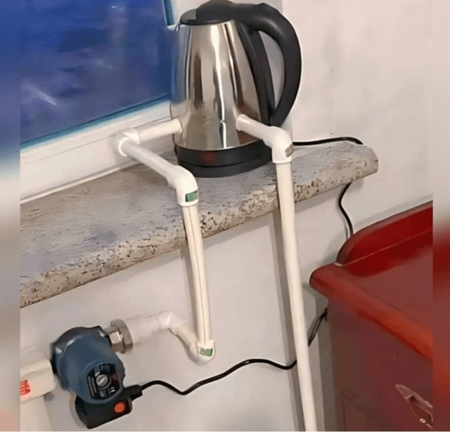 A photograph of a resourceful DIY setup in a studio apartment, presumably crafted by a first-year engineering student. The scene centers on a marble-like countertop holding a stainless steel electric kettle. Below the counter, white PVC pipes form a simple framework, likely part of a homemade water circulation system. A small blue electric pump with a power cord is mounted on the wall, connected to the PVC pipes, suggesting it drives water flow. To the right, a red plastic container is partially visible, likely for water storage. The background includes a window with a blue tint, typical of an apartment setting. This setup exemplifies student ingenuity, using affordable materials to solve a practical need.