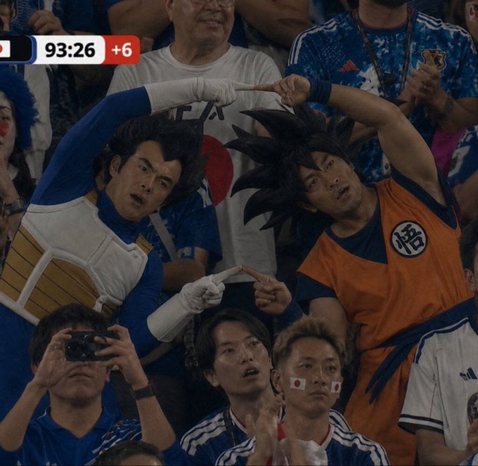 This photograph captures a scene in the spectator stands during a FIFA World Cup match featuring Japanese supporters. At the center, two male fans are cosplaying as iconic Dragon Ball characters Vegeta and Son Goku, performing the famous fusion dance pose by pointing their index fingers toward each other, with engaged, surprised expressions as they watch the match. The match timer visible at the top left shows 93:26 of game time with 6 minutes of stoppage time added. Surrounding the cosplayers are other Japanese fans wearing the national team's blue jerseys, some with Japanese flag decals on their faces, and one fan in the foreground is taking a photo with a camera.