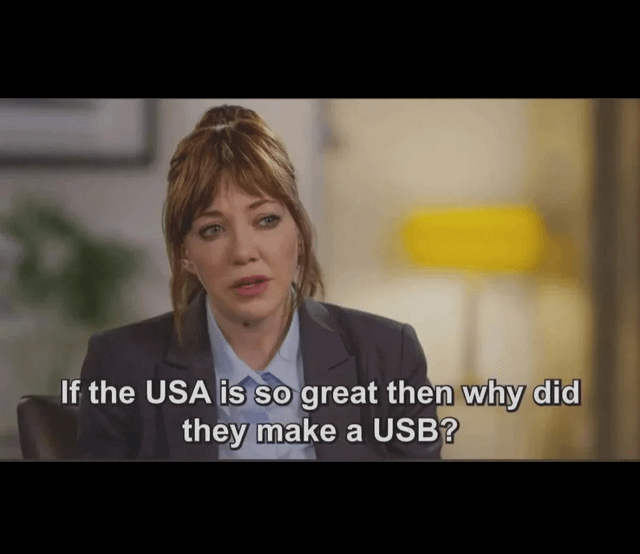 A meme featuring a screenshot of a woman with light brown hair tied in a ponytail, wearing a dark blazer over a light blue shirt, sitting in an indoor setting. The background includes a blurred yellow lamp and a plant. Overlaid text reads: 'If the USA is so great then why did they make a USB?' The humor relies on a pun between 'USA' (United States of America) and 'USB' (Universal Serial Bus), presenting a nonsensical causal relationship for comedic effect.