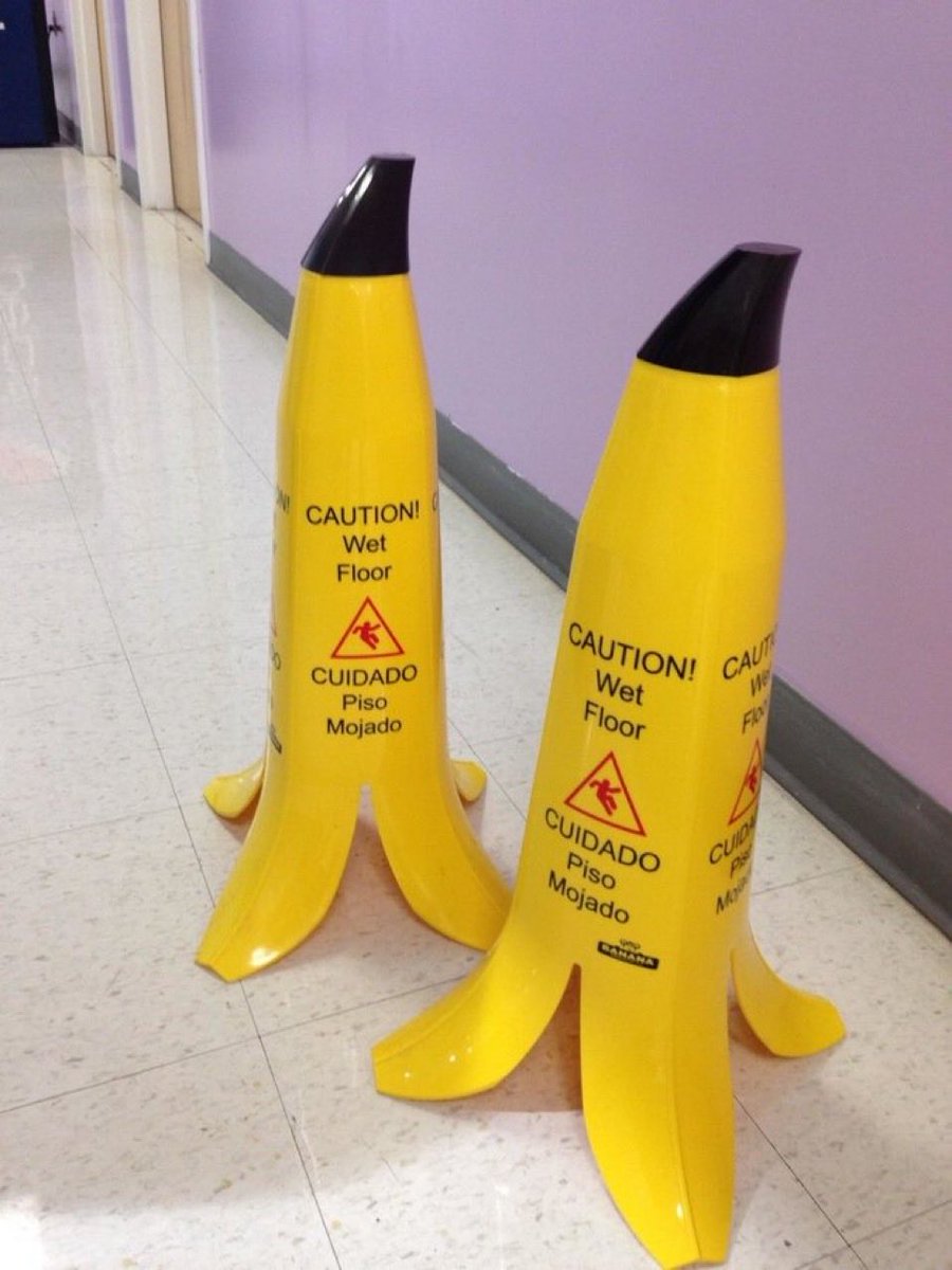 The image displays two freestanding wet floor warning signs creatively designed to look like peeled bananas, placed on a light-colored tiled hallway next to a pale purple interior wall. The bright yellow signs have black tops mimicking banana stems, with four splayed banana peel segments forming the stable base of each sign. Each sign features bilingual safety text: the English line reads "CAUTION! Wet Floor" above a standard red triangular slipping hazard icon, while the Spanish line below reads "CUIDADO Piso Mojado". A small brand logo reading "BANANA" is visible at the bottom of the right sign. The visual gag plays on the classic slapstick comedy trope of people slipping on discarded banana peels, turning the familiar joke into a functional, humorous public safety item.