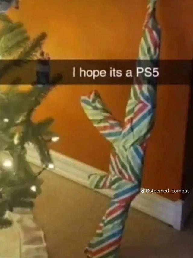 This is a Christmas-themed meme image. On the left, a partially visible decorated Christmas tree with lit lights and ornaments sits against an orange wall. To the right, a gift wrapped in colorful red, green, blue, and white striped paper is positioned to create an optical illusion: its shape and angle make it appear as if it's a person in a dynamic, stretching pose with one arm raised. Overlaid across the upper middle of the image is the text "I hope its a PS5". The humor comes from the contrast between the excited wish for a sleek, popular PS5 gaming console and the misleading, human-like shape of the wrapped gift, playing on the common anticipation of desirable holiday gifts, especially high-demand gaming hardware.