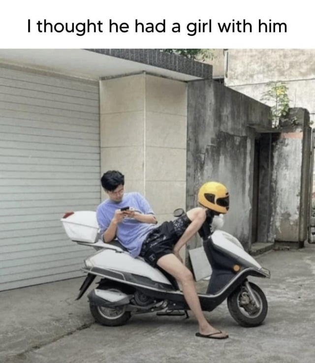 A photograph showing a man sitting on a white scooter parked in an alleyway. The man, wearing a light purple shirt and black shorts, is focused on his phone. The scooter's front has a yellow helmet positioned upright, creating an optical illusion that appears to be a person with a helmet sitting in front of the man. Above the image, there is text that reads 'I thought he had a girl with him,' referencing the illusion of a second person due to the helmet's placement.