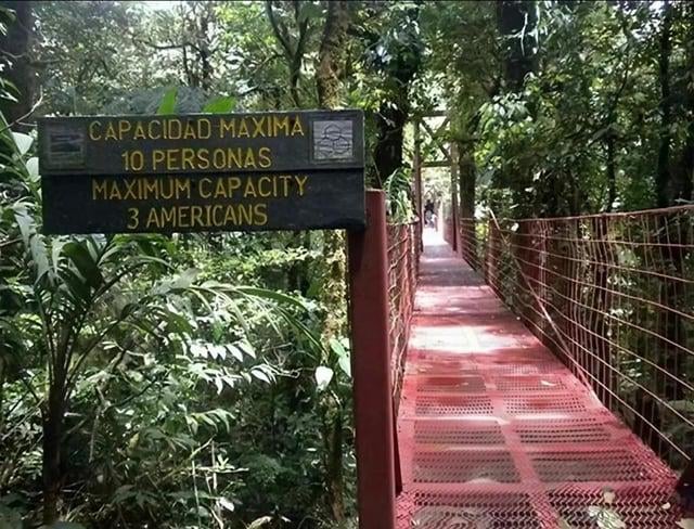 The image depicts a red suspension bridge in a dense forest with a prominent capacity sign. The sign features Spanish text 'CAPACIDAD MAXIMA 10 PERSONAS' (Maximum Capacity 10 People) and below it, an English translation that humorously reads 'MAXIMUM CAPACITY 3 AMERICANS'. This creates a comedic effect by implying a stereotype that Americans occupy more space, leading to a drastically reduced capacity. The bridge has a red metal grid surface and railings, with a few indistinct people visible in the distance walking along it, surrounded by lush green trees and plants.