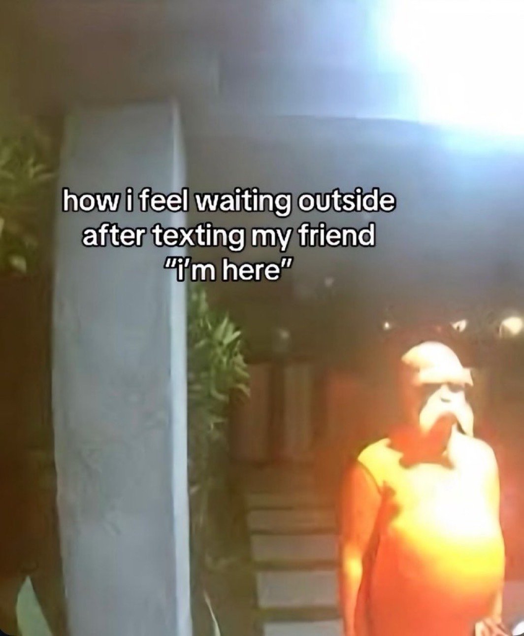 This meme uses a shot of the iconic eerie giant animatronic doll from the first episode of the Netflix series Squid Game, standing motionless outside in dim orange lighting. Overlaid on the upper half of the image is text that reads 'how i feel waiting outside after texting my friend "i'm here"'. The joke draws a humorous relatable comparison between the awkward, uncomfortable feeling of standing around alone waiting for a friend who takes a long time to come meet you after you notify them you have arrived, and the unsettling stationary pose of the Squid Game doll.