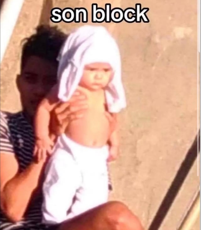 This meme features a low-resolution candid photo taken outdoors on a sunny day, showing an adult in a striped shirt sitting on concrete and holding a young baby. The baby has a white cloth draped over its head resembling a hood, and is positioned directly in front of the adult's face, fully blocking the adult from direct sunlight. Text at the top of the image reads 'son block', a homophonic pun on the common term 'sun block' (sunscreen). The meme's official title 'Total Eclipse of the Son' is a secondary pun, referencing the popular song 'Total Eclipse of the Heart' to joke that the child is acting as a human sun shield and solar eclipse for the parent holding them.