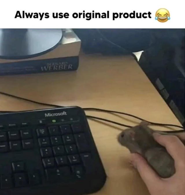 A meme image depicting a tech workspace with a black Microsoft keyboard on a wooden desk. A human hand is shown holding a real live mouse (the animal) in place of a computer mouse, playing on the phrase 'Always use original product 😂' displayed at the top. In the background, there is a book titled 'The Elder Scrolls IV: Oblivion' and a computer monitor. The humor arises from the literal interpretation of 'original product'—using the biological/original mouse instead of an electronic computer mouse.