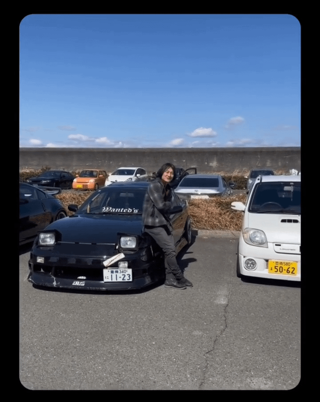 The image depicts a real man with a smiling, friendly expression leaning against the driver's side door of a modified black Japanese sports car, likely a Nissan Silvia model, in an outdoor parking lot under a clear, bright blue sky. The car features a 'Wanted's' decal on its windshield, a white temporary-style license plate with the number '1-23' and Japanese text above it, plus aftermarket front bumper and headlight components. To the right of the black car sits a small white kei car with a yellow Japanese license plate displaying '50-62'. In the background, multiple other vehicles—including an orange kei car, black sports cars, and sedans—are parked near dry, brown grass and a tall concrete barrier. The scene suggests a casual car meet or gathering for automotive enthusiasts, set in Japan, as evidenced by the vehicle styles and Japanese license plates.