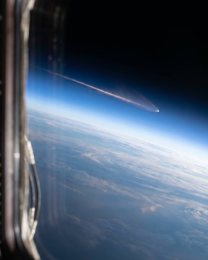 This is a photograph captured from a window on the International Space Station (ISS), showing the Artemis mission capsule during its atmospheric re-entry above Earth. A bright glowing contrail stretches from the capsule across the view of space and atmosphere. The edge of the ISS window frames the shot on the left, while below is the curved cloud-covered surface of Earth transitioning from the blue atmospheric layer to the blackness of outer space in the upper right.