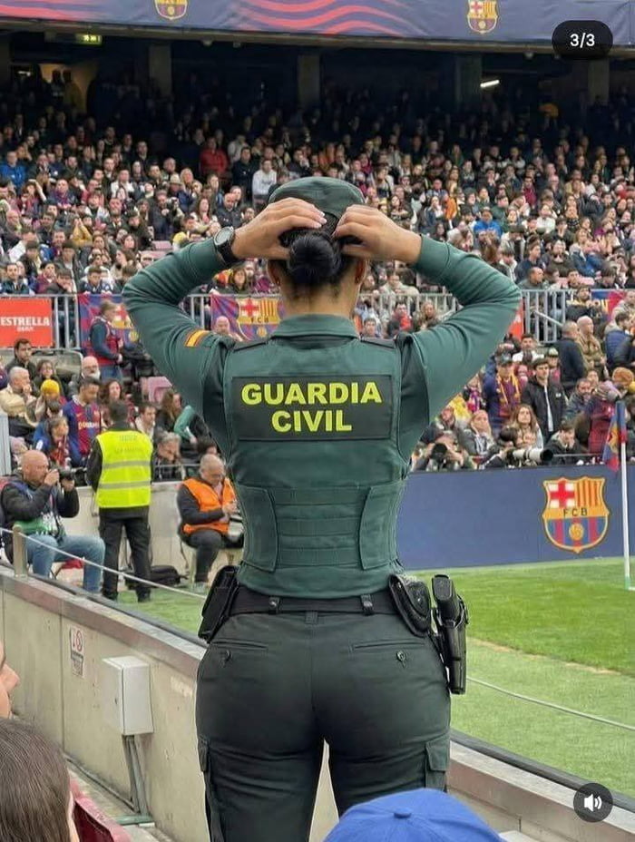 A photograph of a female Guardia Civil officer standing in front of a crowded football stadium, likely Camp Nou (home of FC Barcelona). She faces away from the camera, adjusting her hair with both hands behind her head. Her dark green uniform has 'GUARDIA CIVIL' in bold yellow letters on the back. The background features spectators, many in FC Barcelona colors, with the club's logo visible on the stadium barrier. The original title 'There's nothing civil here' plays on the double meaning of 'civil'—referencing the Guardia Civil and potentially the lively, uncivil atmosphere of a sports crowd.