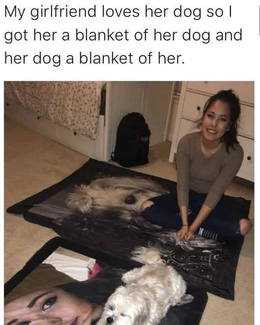 The meme has a text caption above a real-life photograph. The caption reads 'My girlfriend loves her dog so I got her a blanket of her dog and her dog a blanket of her.' The photo shows a young woman smiling happily as she sits cross-legged on a large custom blanket printed with a photo of her small, light-colored fluffy dog. In front of her, the actual dog rests comfortably on a second custom blanket printed with a photo of the woman. The scene is set in a carpeted bedroom, with a dresser and backpack visible in the background, showcasing a thoughtful, endearing romantic gesture that pleases both the girlfriend and her pet.