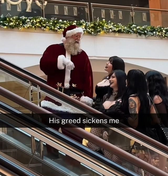 A photograph taken in a shopping mall during the holiday season. Santa Claus, dressed in his traditional red suit with white trim and black belt, stands near an escalator. He is interacting with a group of four women who have long black hair, wear gothic-style clothing (including black dresses, fishnets, chokers, and visible tattoos). The women appear engaged in conversation with Santa, some smiling. The mall background features festive decorations like garlands with white lights, indicating a Christmas setting. At the bottom of the image, a text overlay reads: 'His greed sickens me'.