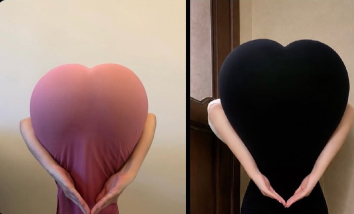 This is a side-by-side optical illusion image featuring two people posing to create a clever visual trick. At first glance, most viewers think each person is holding a large heart-shaped pillow, but the illusion is actually created by the person's pose and matching solid-colored clothing: their bodies form the heart shape, with their arms curving around the outside to complete the look, tricking the eye into seeing a separate pillow. The visual plays on the romantic phrase 'My heart belongs to you', with the heart shape being the focal point. The left panel uses a pink dress against a plain light wall, while the right panel uses a black dress against a wall next to a wooden door frame.