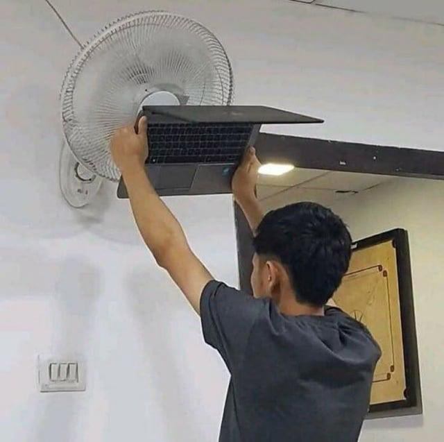 This meme depicts a young man standing and holding his open laptop directly pressed against the spinning grates of a wall-mounted ceiling fan, as an extremely exaggerated, absurd solution for cooling an overheating device. The accompanying title references the well-known issue of the video editing software DaVinci Resolve being extremely resource-heavy, which commonly causes laptops to overheat drastically even after just 15 minutes of use, leading users to go to ridiculous lengths to cool their devices while editing.