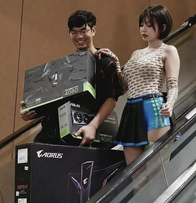 The image depicts a young man wearing glasses, grinning widely with visible excitement, standing on an escalator while holding three boxes of premium PC hardware: two GeForce RTX 4080 graphics cards (one from Galax, one from ASUS) and a large AORUS branded PC case box. A woman stands next to him, resting one hand on his shoulder with an unimpressed, neutral expression. The humor comes from the widespread understanding among PC building and gaming communities that high-end flagship graphics cards like the RTX 4080 are very expensive and highly desirable, making the man's excitement at acquiring multiple of these products relatable to tech enthusiasts, hence the joke that he is the 'luckiest person'.