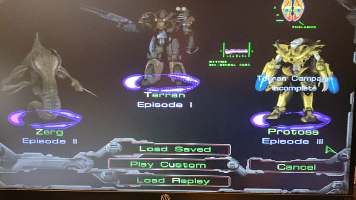 A screenshot of a classic video game's episode selection menu, likely from StarCraft. The dark background features three faction-specific episode icons: Zerg Episode II (left, purple ring), Terran Episode I (center, purple ring), and Protoss Episode III (right, purple ring with 'Terran Campaign incomplete' text overlay). Below the icons are four UI buttons: 'Load Saved', 'Play Custom', 'Load Replay', and 'Cancel'. A small HP monitor logo is visible at the bottom center of the screen.