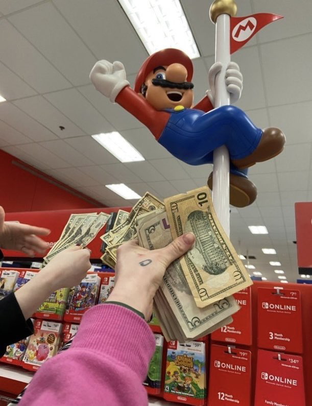 This is a humorous photograph taken inside a US retail store, likely Target identifiable by its signature red interior decor. A large official plastic Super Mario figure is posed sliding down his signature M-branded flagpole above a display of Nintendo Switch products, including physical game cases such as Animal Crossing and Nintendo Online membership subscription cards. Multiple people are holding out thick stacks of US dollar bills towards the Mario figure, as if offering to pay the character directly. The joke satirizes the high cost of Nintendo products and the large sums of money consumers regularly spend on Nintendo games, subscriptions, and merchandise.