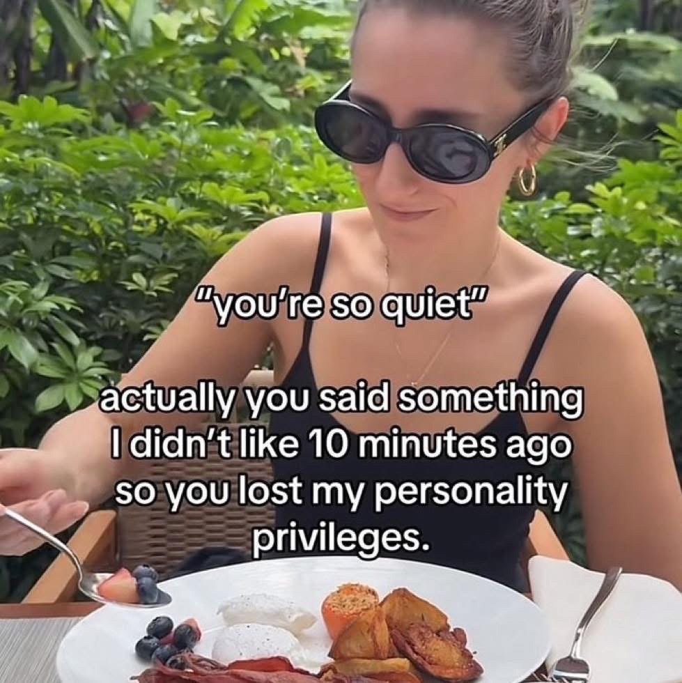 The image features a woman wearing black oval sunglasses, a black camisole, and gold hoop earrings, sitting outdoors at a dining table surrounded by lush green foliage. In front of her is a plate of breakfast including poached eggs, roasted potatoes, bacon, and fresh berries, and she is holding a spoon with cut fruit on it, wearing a subtle, smug unimpressed smirk. Overlaid text first quotes another person saying "you're so quiet", followed by an explanation that the quiet person is reserved because the other person said something they disliked 10 minutes earlier, so the other person has lost access to the quiet person's personality. The meme jokes about the common relatable behavior of withdrawing and being quiet around people who have annoyed you, even if they do not realize they did something wrong.