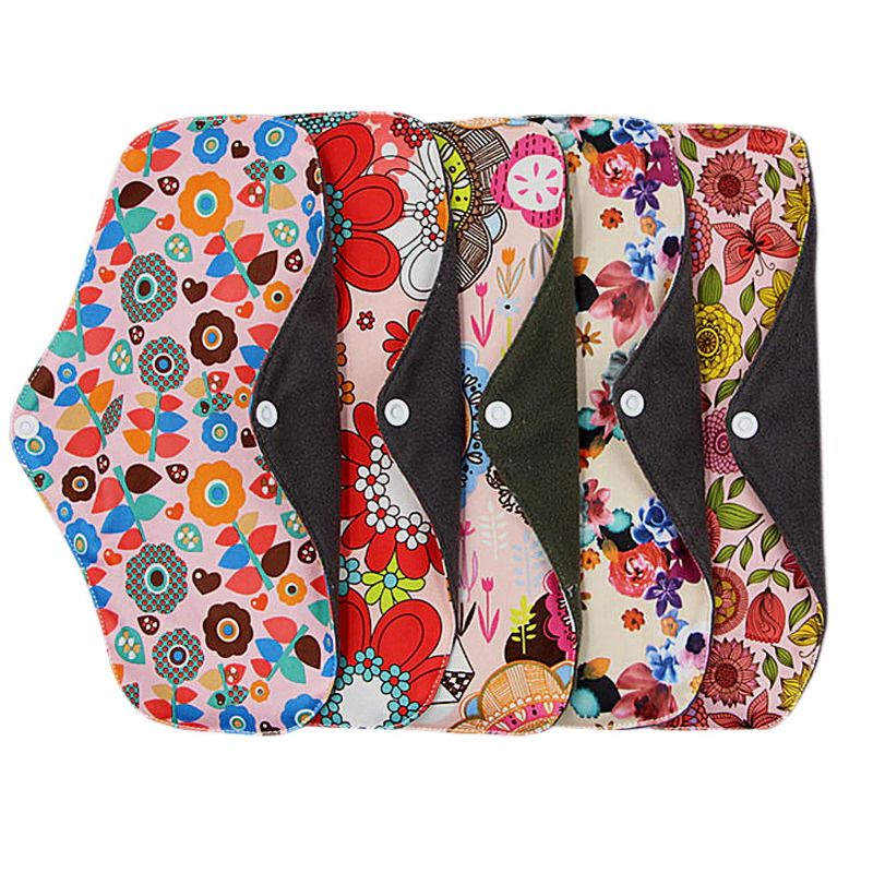 The image shows five reusable cloth menstrual pads arranged horizontally against a plain white background. Each pad has a unique, vibrant floral-patterned outer layer, with designs including retro stylized flowers, watercolor-style florals, and bohemian floral motifs. All pads feature a dark charcoal or dark green absorbent inner lining, and a white plastic snap button on each wing to fasten the pad securely to underwear. These products are marketed as an eco-friendly, zero-waste, reusable alternative to disposable menstrual hygiene products.