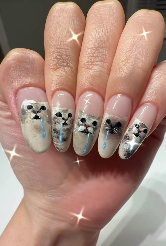 The image displays a person's outstretched hand, featuring long almond-shaped acrylic nails with a sheer nude glossy base. Each nail has hand-painted artwork of the iconic crying/snotty cat reaction meme: each cat face has wide dark eyes, an open mouth, and a single shiny bright blue drip resembling snot or tears coming from the nose area. Sparkle filter effects are scattered across the hand and nails to emphasize the glossy finish of the manicure.