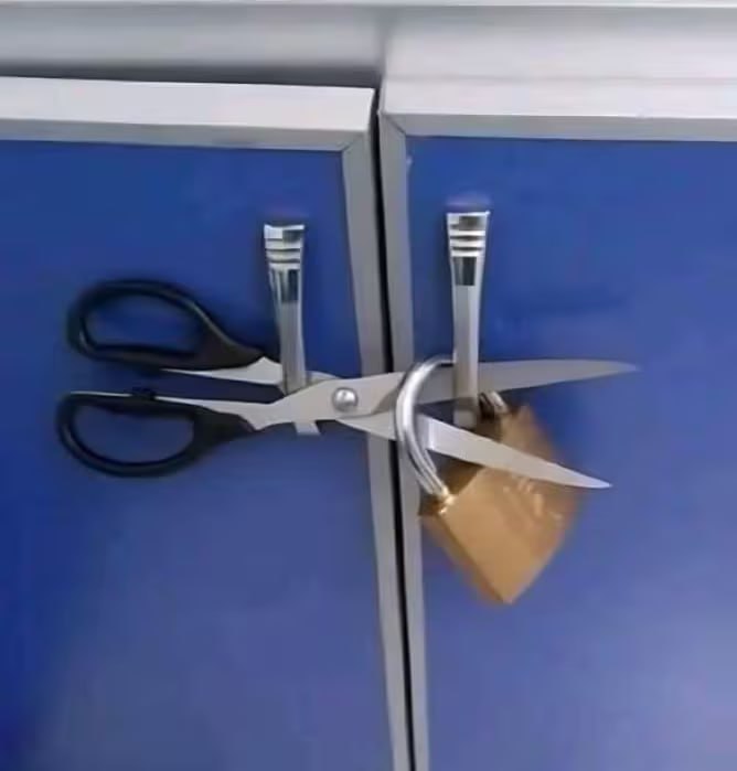 A photograph showing two adjacent blue cabinet doors with metallic vertical handles. A pair of silver scissors with black handles is horizontally wedged between the two handles, with the blades crossing over the right handle. A brass padlock is attached to the scissors' blades, using the scissors as an improvised lock to secure the cabinet doors shut. The setup is an unconventional and creative solution for locking the cabinets without a traditional lock mechanism.