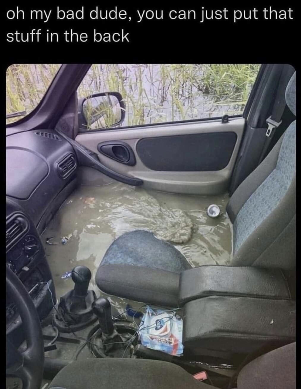 The image displays the interior of a passenger car that is heavily flooded, with murky, dirty brown water completely filling the entire footwell area, submerging the lower portions of the seats, manual gear shifters, and small loose items including a soda can and a wet wipe package scattered on the floor. Outside the car windows, tall reeds and open water are visible, indicating the vehicle is partially submerged in a body of water. The text at the top of the meme reads 'oh my bad dude, you can just put that stuff in the back', creating a humorous, absurd joke as the request is completely impractical: there is no dry space in the car to place any items, making the polite, casual request hilariously out of touch with the actual state of the vehicle.