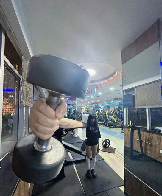This image is taken in an indoor gym, using a forced perspective optical illusion to create a humorous visual effect. A person holds a dumbbell extremely close to the camera, making their hand and the dumbbell appear disproportionately massive, as if the giant hand is about to grab the woman standing at a distance in the background of the gym. The background shows standard gym equipment, exercise mats, large mirrors, ceiling lights, and windows overlooking a nighttime street scene.