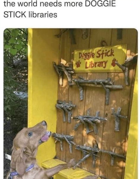 A photograph showing a happy golden-brown dog standing on its hind legs with front paws resting on a small yellow wooden structure labeled 'Doggie Stick Library' (decorated with red paw print symbols). The library features multiple sticks arranged on its wooden panels, and the dog appears excited, looking up at the sticks with its mouth open and tongue out. Above the image, prominent text reads 'the world needs more DOGGIE STICK libraries', emphasizing the playful and wholesome concept of a community resource for dogs.