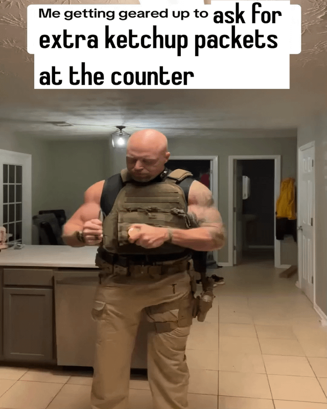 A meme image featuring a real man standing in a kitchen, dressed in tactical gear including a military-style vest, utility pants, and a holster. He is in the process of adjusting his vest with a focused expression. The top of the image contains a white caption box with black text that reads: 'Me getting geared up to ask for extra ketchup packets at the counter'. The humor stems from the夸张 contrast between the intense, military-like preparation and the mundane, low-stakes social task of requesting ketchup, satirizing the anxiety introverts may feel about simple social interactions.