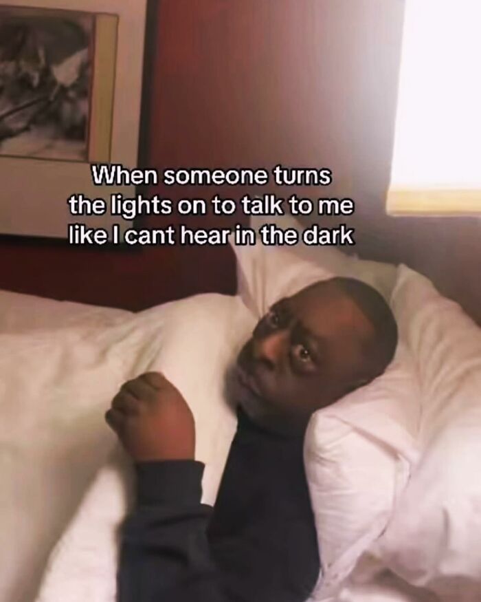 This single-panel reaction meme features a Black man lying in bed under white bedding, looking visibly annoyed and squinting as a bright light is suddenly turned on next to him. Overlaid text above him reads 'When someone turns the lights on to talk to me like I cant hear in the dark'. The joke is based on the relatable, frustrating experience of being woken up or disturbed while resting, when the person trying to speak to you unnecessarily turns on the lights despite the fact that hearing works perfectly in the dark, causing an unnecessary unpleasant interruption.