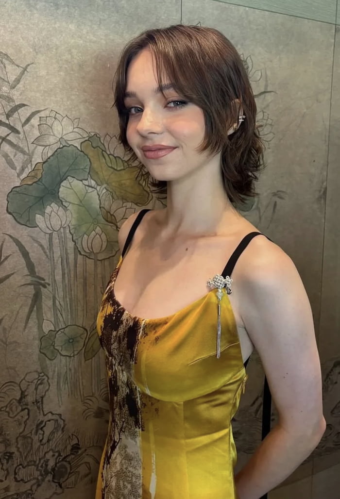 A photograph of a woman with short brown hair wearing a yellow silk dress featuring black strap details and a decorative brooch on the shoulder. She stands against a backdrop of traditional Chinese-style artwork depicting green lotus leaves and flowers. The woman smiles softly, with one arm positioned behind her back, creating an elegant and composed pose.