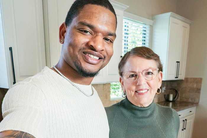 A color photograph capturing two individuals taking a selfie in a kitchen environment. The man on the left is dressed in a light-colored ribbed sweater, accessorized with a silver necklace, and has a visible tattoo on his left arm. He is smiling broadly, likely holding the camera to take the selfie. To his right stands an older woman wearing a green turtleneck sweater, glasses, and hoop earrings, also smiling warmly. The kitchen background features white upper cabinets, a window with horizontal blinds allowing natural light, and a countertop with a small metallic appliance, contributing to a cozy, domestic atmosphere.