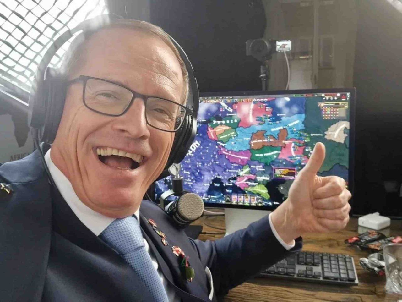 The image features a man wearing a dark suit, white shirt, and blue tie, with black headphones over his ears. He is smiling widely and giving a thumbs-up gesture with his right hand. Behind him, a large monitor displays a colorful, detailed map with various labeled regions and icons, resembling the interface of a strategy video game (e.g., Hearts of Iron or Europa Universalis). A microphone is positioned in front of him, suggesting a broadcasting or streaming setup. The background includes a window with a grid-patterned covering and some studio equipment, indicating a professional or semi-professional recording environment.