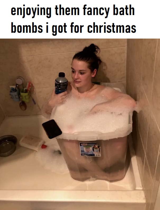 A humorous image depicting a person sitting in a plastic storage bin placed inside a bathtub, filled with copious bath bubbles. The individual is holding a bottle in one hand, with a phone resting on the edge of the bin. Above the image, text reads: 'enjoying them fancy bath bombs i got for christmas,' highlighting the comedic contrast between the 'fancy' bath bombs and the improvised use of a storage bin as a bath container.