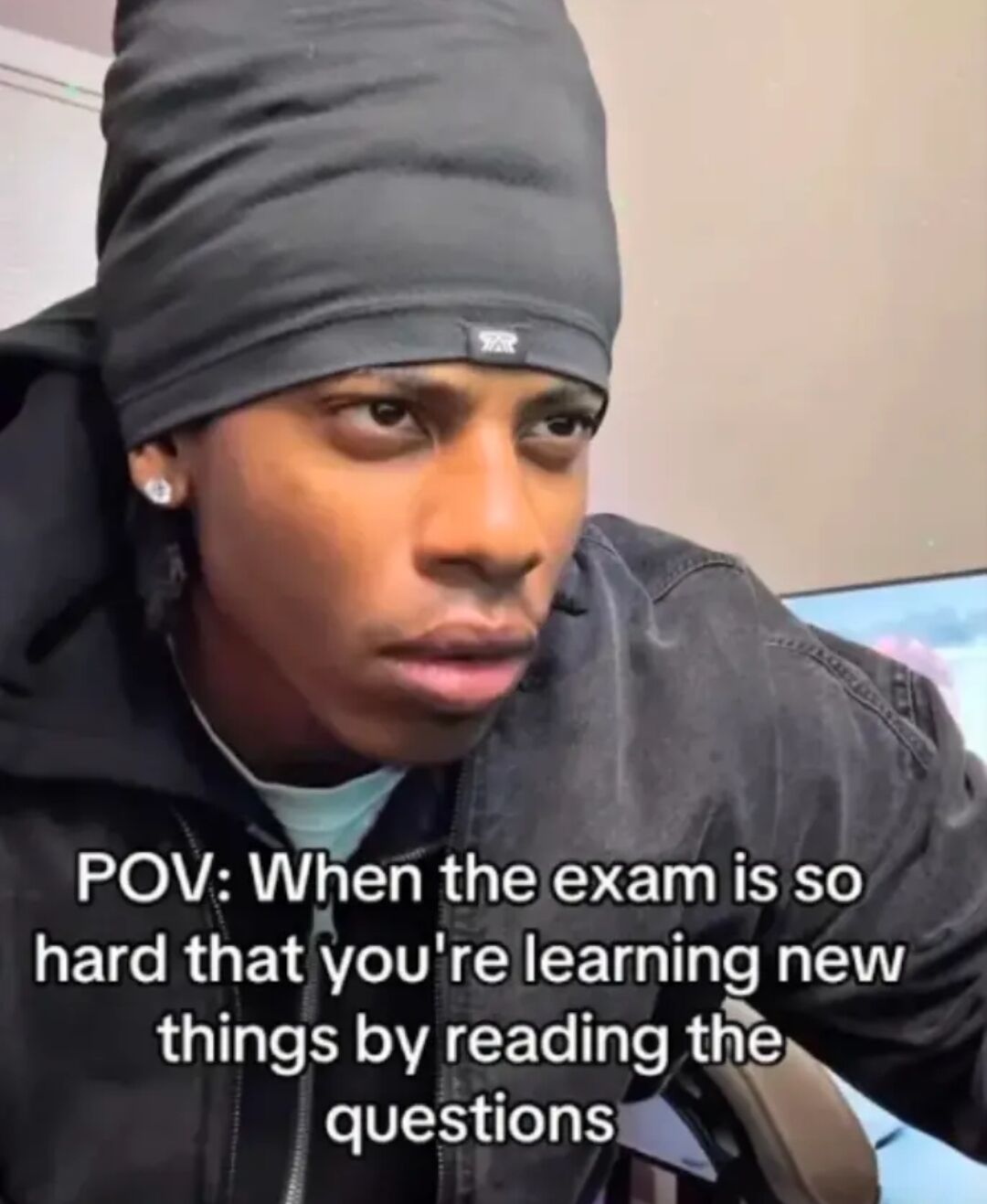 A meme featuring a real person wearing a black beanie and dark hoodie, leaning forward with a surprised or confused expression. Overlaid text reads 'POV: When the exam is so hard that you're learning new things by reading the questions,' humorously depicting the frustration and relatable experience of encountering unexpectedly difficult exam questions that introduce new information not previously studied.
