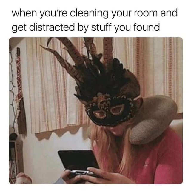 This is a single-panel relatable meme. The top of the meme has text that reads 'when you're cleaning your room and get distracted by stuff you found'. Below the text is a photograph of a blonde person wearing an ornate black feathered masquerade mask, with a plush curved ram's horn accessory resting on their shoulder, looking down with focused attention at an old flip phone they are holding in both hands. The joke conveys the extremely common relatable experience of starting to clean one's living space, stumbling across forgotten old random items, and becoming fully sidetracked from the chore to interact with or use those found items instead.